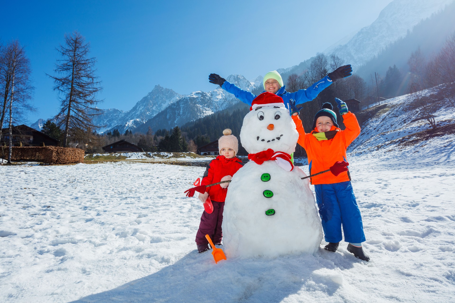 Three kids play with snowman lifting hands stand over mountains